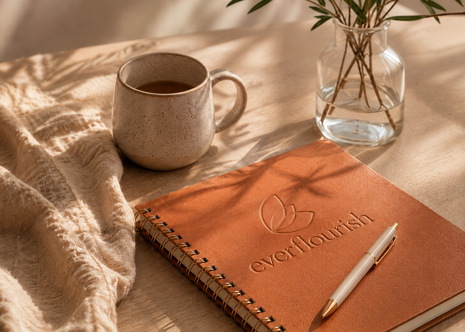 A terracotta notebook embossed with flourish, a cream pen, a speckled mug, a small vase with greenery, and a soft knit blanket in warm window light